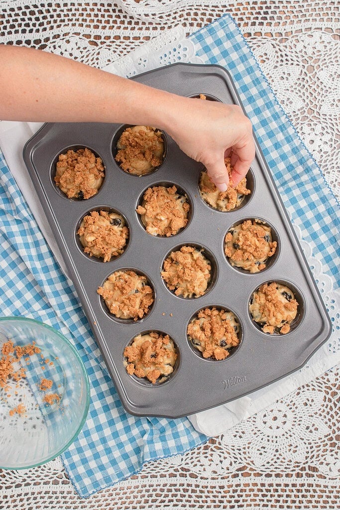 Overhead view of a hand sprinkling cinnamon brown sugar streusel topping onto unbaked blueberry muffins in a gray Wilton muffin tin, with a small glass bowl of remaining streusel on a blue gingham cloth and white lace doily.
