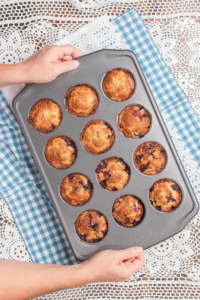 Overhead view of hands holding a gray Wilton muffin tin filled with freshly baked golden blueberry muffins made with freshly milled flour, set on a blue gingham cloth and white lace doily.