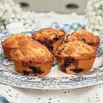 Four bakery-style blueberry streusel muffins made with freshly milled flour arranged on a blue and white transferware plate, with one muffin broken open to show the tender crumb and juicy blueberries, surrounded by white baby's breath flowers with a cottage kitchen sink in the background.