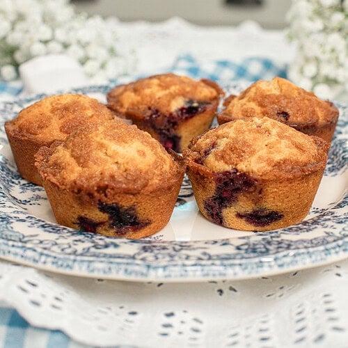 Four bakery-style blueberry streusel muffins made with freshly milled flour arranged on a blue and white transferware plate, with one muffin broken open to show the tender crumb and juicy blueberries, surrounded by white baby's breath flowers with a cottage kitchen sink in the background.