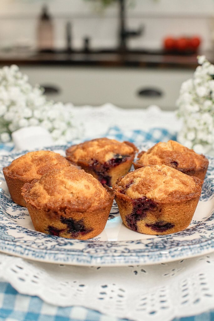 Four bakery-style blueberry streusel muffins made with freshly milled flour arranged on a blue and white transferware plate, with one muffin broken open to show the tender crumb and juicy blueberries, surrounded by white baby's breath flowers with a cottage kitchen sink in the background.