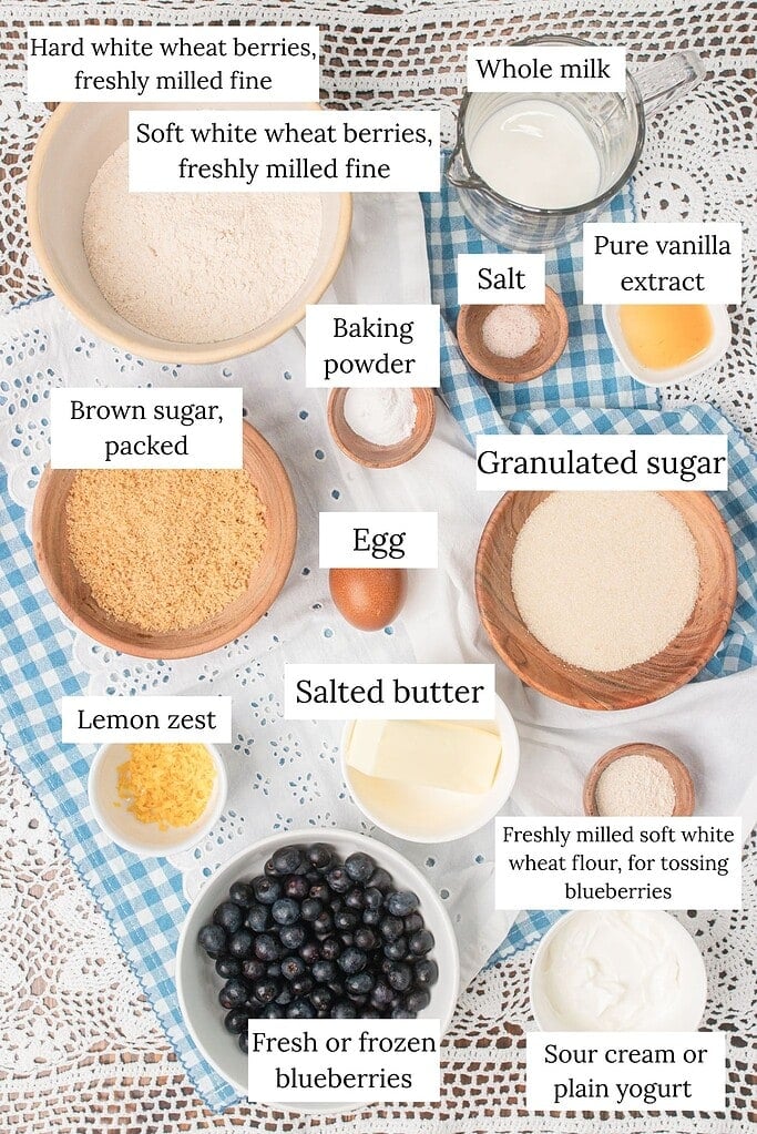 Overhead flat lay of blueberry streusel muffin ingredients on a blue gingham cloth and white lace doily, including bowls of hard white wheat and soft white wheat freshly milled flour, brown sugar, granulated sugar, whole milk, egg, salted butter, lemon zest, fresh blueberries, sour cream, baking powder, salt, and pure vanilla extract.