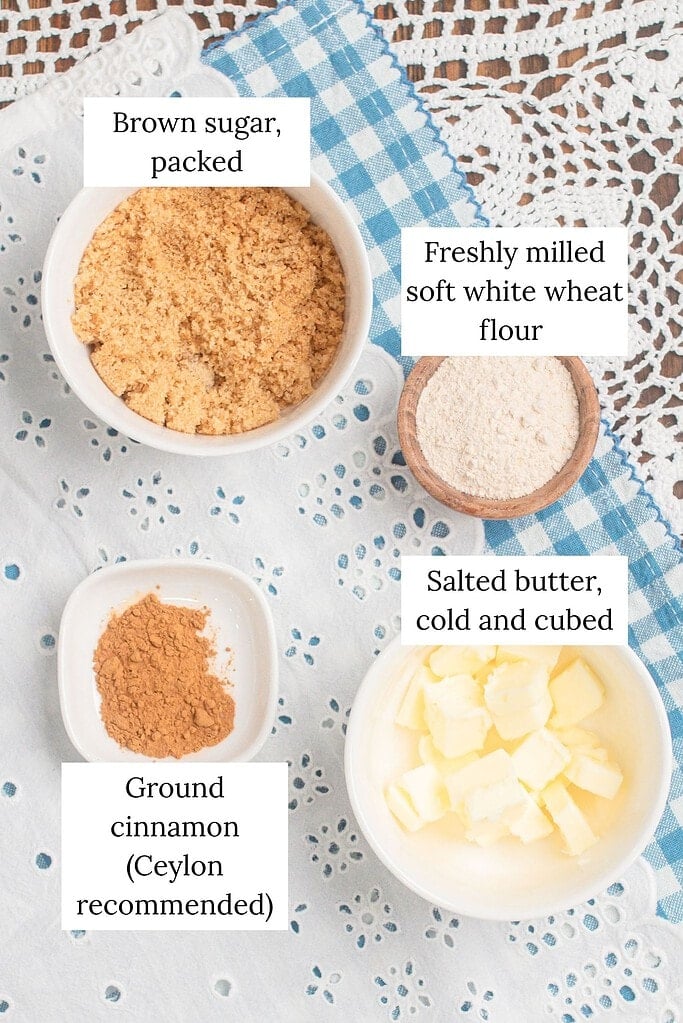 Overhead flat lay of cinnamon streusel topping ingredients on a blue gingham cloth and white eyelet linen, including bowls of packed brown sugar, freshly milled soft white wheat flour, ground cinnamon, and cold cubed salted butter.