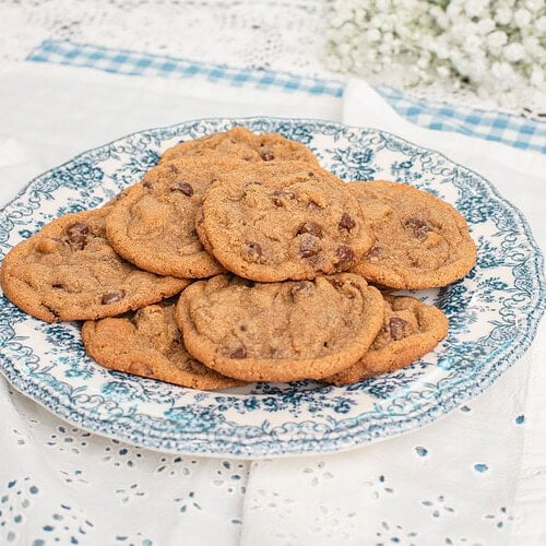 A blue and white floral plate piled with brown butter chocolate chip cookies made with freshly milled flour on a white eyelet tablecloth with baby's breath flowers and sage green cabinets in the background