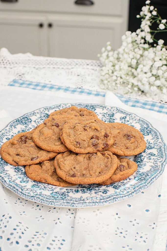 A blue and white floral plate piled with brown butter chocolate chip cookies made with freshly milled flour on a white eyelet tablecloth with baby's breath flowers and sage green cabinets in the background