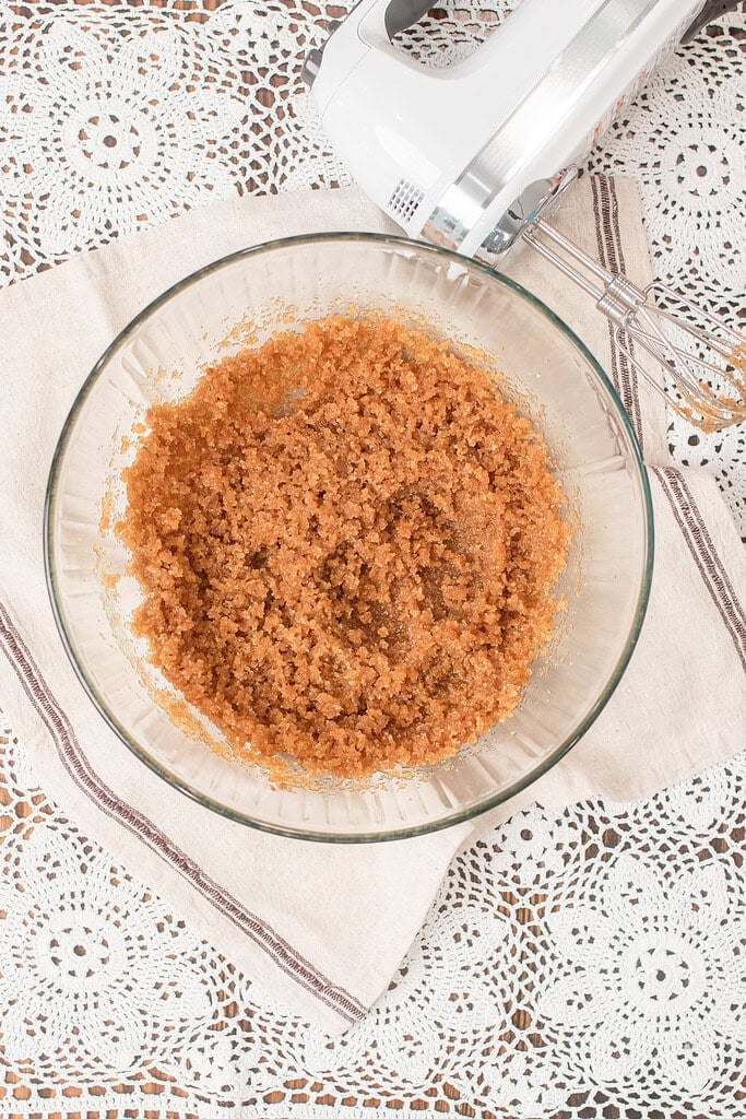 Overhead view of a glass mixing bowl filled with creamed brown butter and sugar for brown butter chocolate chip cookies made with freshly milled flour, with a hand mixer resting on a lace doily beside it