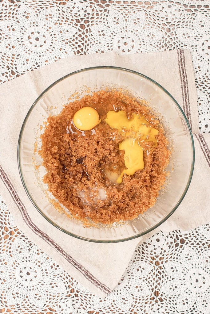 Overhead view of a glass mixing bowl with eggs added to the brown butter and sugar mixture for fresh milled flour chocolate chip cookies, set on a linen napkin and lace doily