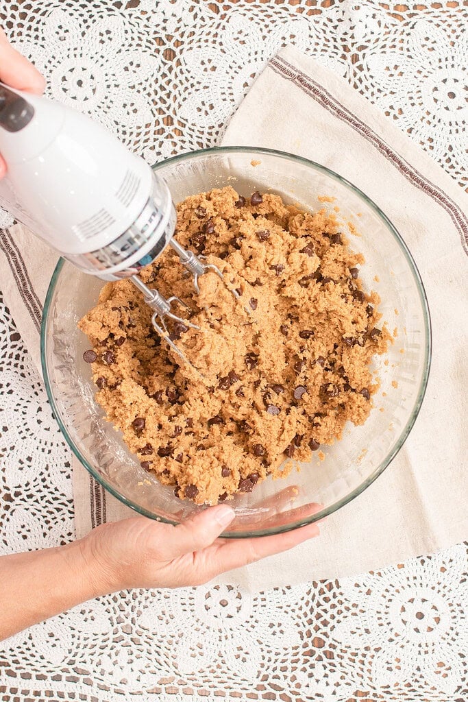 A hand mixer blending freshly milled flour chocolate chip cookie dough loaded with chocolate chips in a glass bowl set on a linen napkin and lace doily