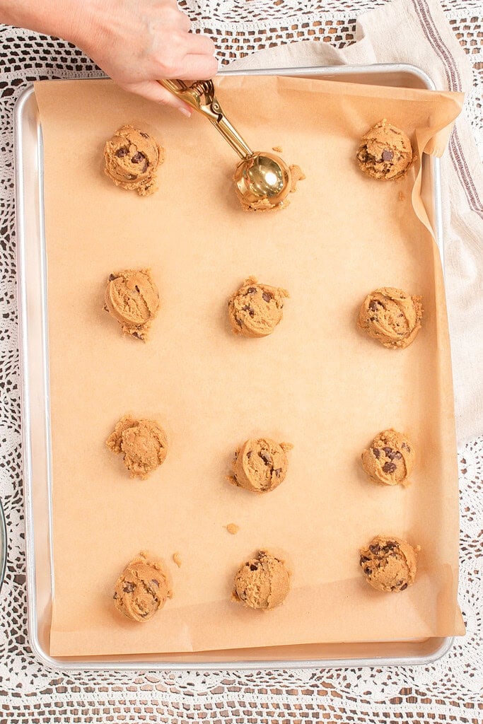A hand using a gold cookie scoop to portion brown butter chocolate chip cookie dough made with freshly milled flour onto a parchment-lined baking sheet