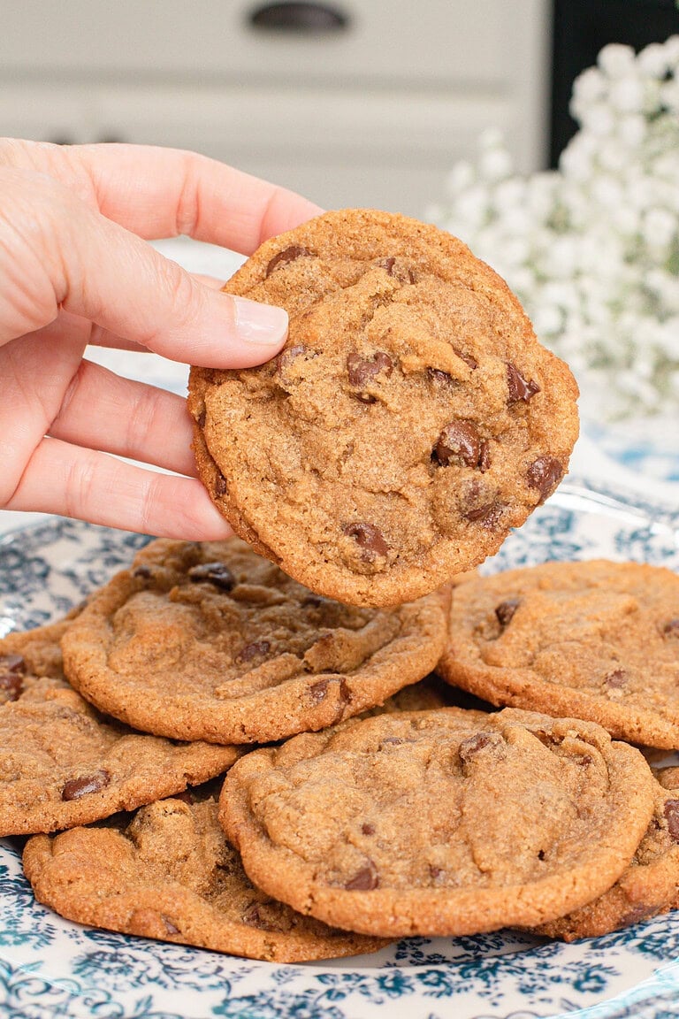 A hand holding a single brown butter chocolate chip cookie made with freshly milled flour up close, showing the golden texture and melted chocolate chips, with more cookies on a blue floral plate in the background