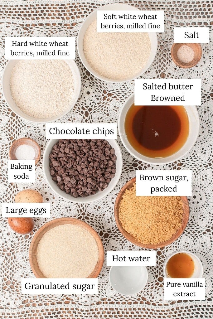 Overhead flatlay of labeled ingredients for brown butter chocolate chip cookies made with freshly milled flour, including hard white and soft white wheat berries milled fine, browned butter, chocolate chips, brown sugar, eggs, vanilla, baking soda, and salt on a lace doily