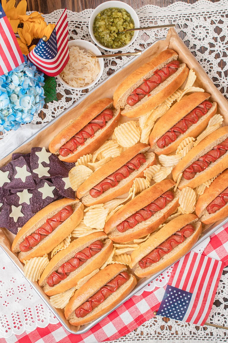 American flag hot dog board with fresh milled flour buns, ridged potato chips, and blue corn star tortilla chips, served with pickle relish and sauerkraut on a lace tablecloth