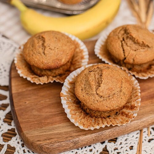 Banana muffins with freshly milled flour cooling on a wood board next to a ripe banana and muffin tin