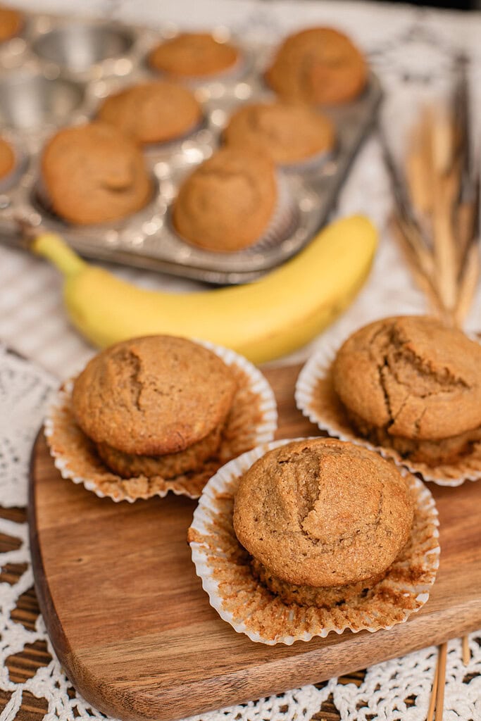 Banana muffins with freshly milled flour cooling on a wood board next to a ripe banana and muffin tin
