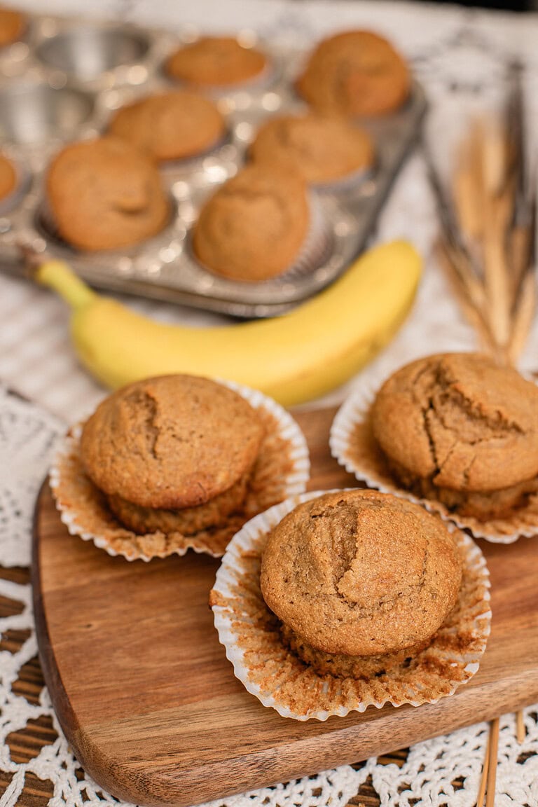 Banana muffins with freshly milled flour cooling on a wood board next to a ripe banana and muffin tin