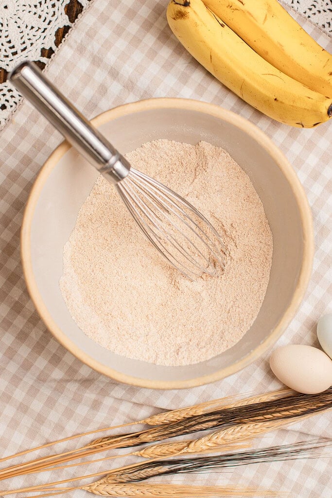 Whisking freshly milled flour in a bowl surrounded by ripe bananas, wheat stalks, and eggs for banana muffins