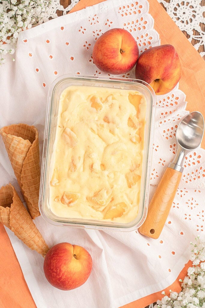 Overhead view of brown butter peach custard ice cream in a glass container with waffle cones and fresh peaches