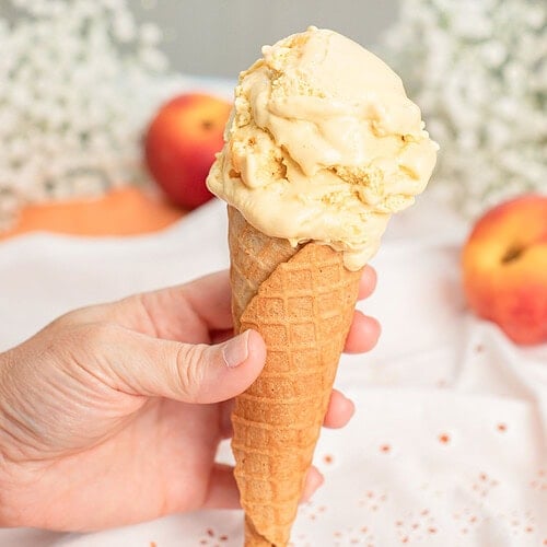 Hand holding a waffle cone topped with a scoop of brown butter peach custard ice cream