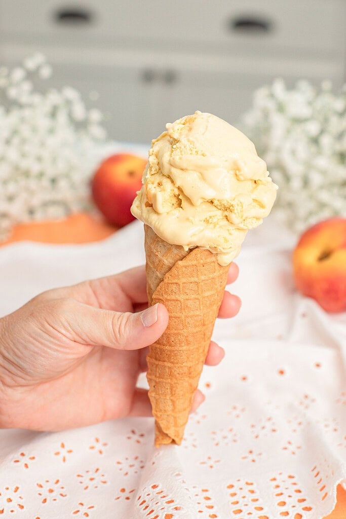 Hand holding a waffle cone topped with a scoop of brown butter peach custard ice cream