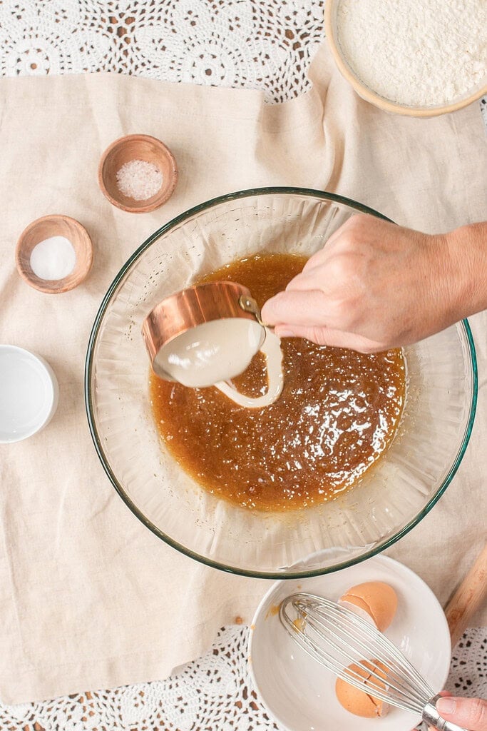 Sourdough discard being poured from a copper measuring cup into the browned butter and egg mixture for chocolate chip cookie dough
