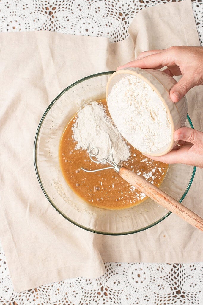 All-purpose flour being poured into the wet brown butter sourdough cookie batter with a wooden spoon resting in the bowl
