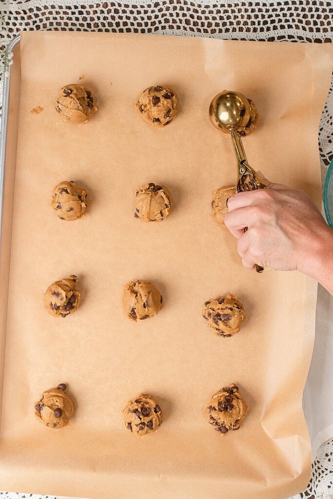 Scoops of brown butter sourdough chocolate chip cookie dough portioned onto a parchment-lined baking sheet with a gold cookie scoop
