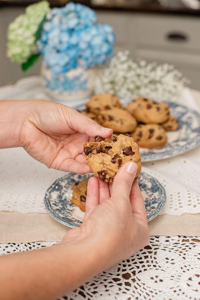 Hands sharing a warm brown butter sourdough chocolate chip cookie broken in half to reveal melted chocolate chips, with blue hydrangeas and cookies on a floral plate in the background