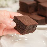 A hand holding a cakey brownie made with freshly milled flour showing its soft fluffy texture, with more brownies blurred on a plate behind