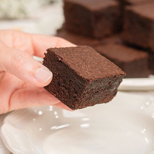 A hand holding a cakey brownie made with freshly milled flour showing its soft fluffy texture, with more brownies blurred on a plate behind