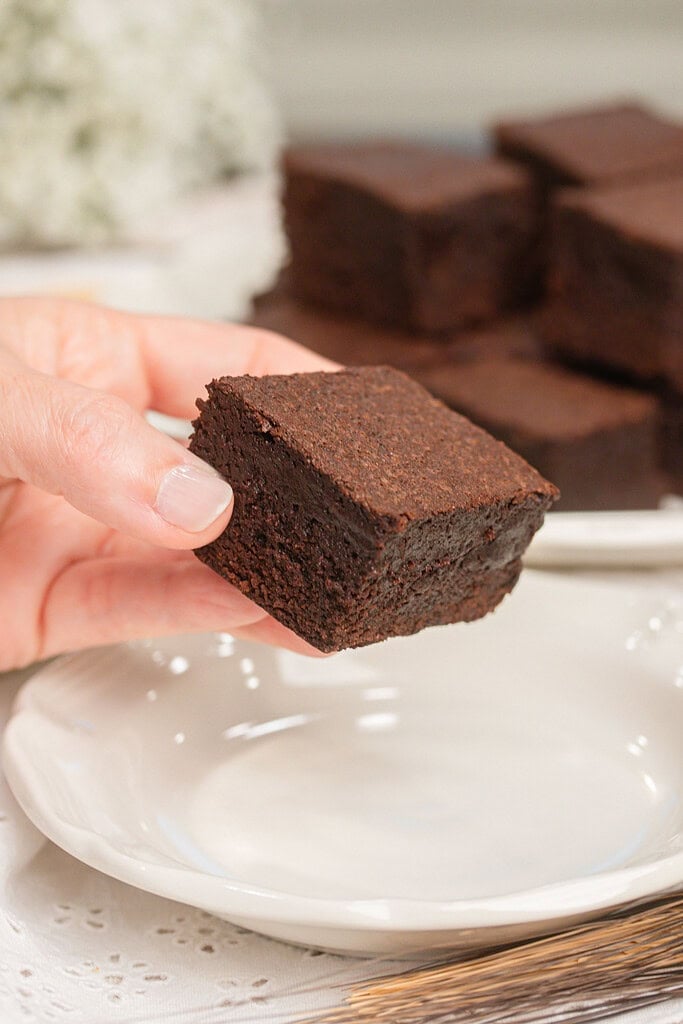 A hand holding a cakey brownie made with freshly milled flour showing its soft fluffy texture, with more brownies blurred on a plate behind