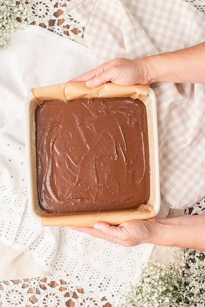 Hands holding a parchment-lined square baking pan filled with unbaked cakey brownie batter made with freshly milled flour