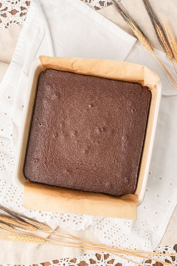 A freshly baked pan of cakey brownies made with freshly milled flour cooling in a parchment-lined baking dish with wheat stalks nearby
