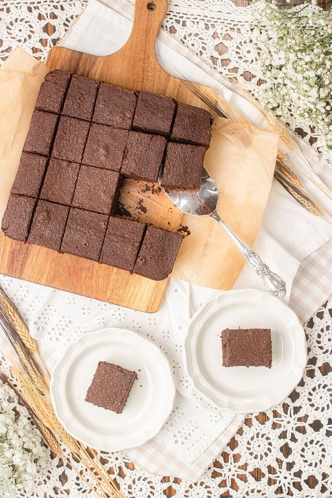 Cakey brownies made with freshly milled flour sliced into squares on a wooden board with one being lifted by a vintage serving spoon and two plated in the foreground