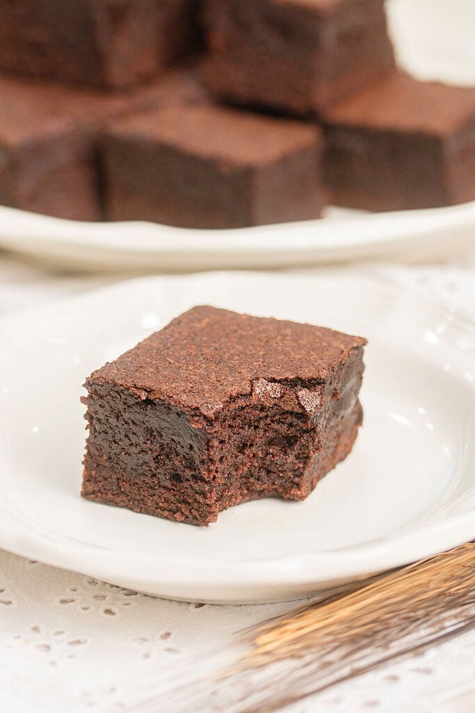 A cakey brownie made with freshly milled flour with a bite taken out showing its soft crumb, with more brownies blurred on a plate behind