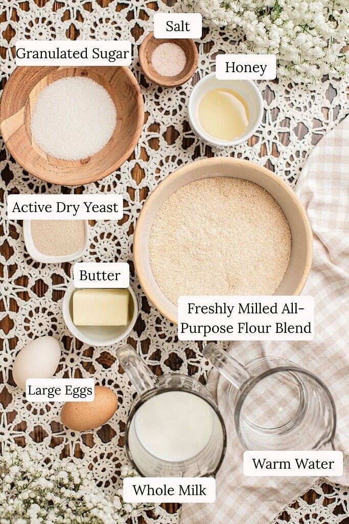 Flat lay of labeled ingredients for dinner rolls made with freshly milled flour, including a bowl of freshly milled all-purpose flour blend, granulated sugar, salt, honey, active dry yeast, butter, large eggs, whole milk, and warm water, arranged on a lace and checked cloth with baby's breath flowers.