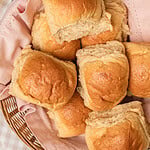 Overhead close-up of soft golden dinner rolls made with freshly milled flour piled in a basket lined with a pink cloth, one roll torn open to show the fluffy interior, with dried wheat stalks resting alongside.