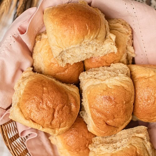 Overhead close-up of soft golden dinner rolls made with freshly milled flour piled in a basket lined with a pink cloth, one roll torn open to show the fluffy interior, with dried wheat stalks resting alongside.