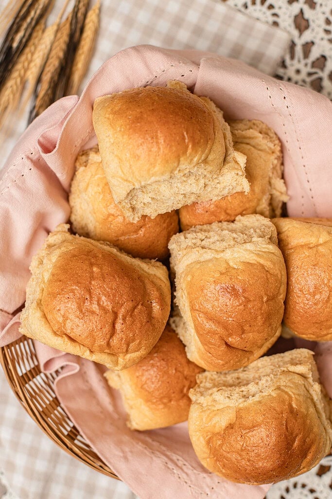 Overhead close-up of soft golden dinner rolls made with freshly milled flour piled in a basket lined with a pink cloth, one roll torn open to show the fluffy interior, with dried wheat stalks resting alongside.