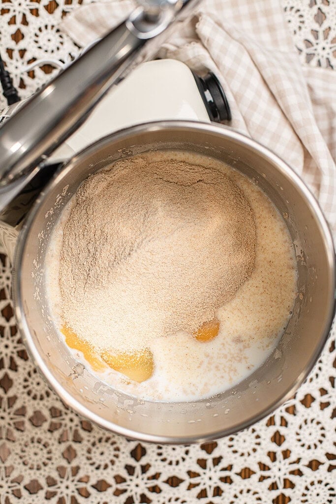 Overhead view of freshly milled flour, milk, and egg yolks added to a stand mixer bowl, not yet mixed together, with the mixer's dough hook visible above.