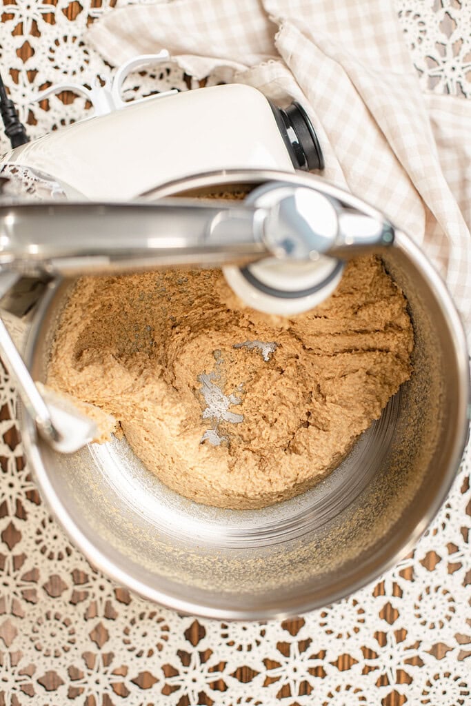 Overhead view of shaggy dinner roll dough being mixed in a stand mixer bowl, not yet smooth, with the dough hook resting in the mixture.