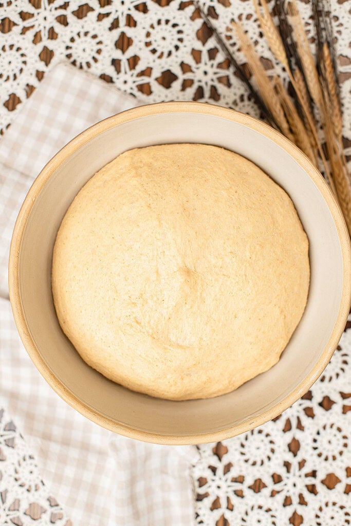 Overhead view of dinner roll dough made with freshly milled flour risen and doubled in a beige bowl, with dried wheat stalks resting beside it.