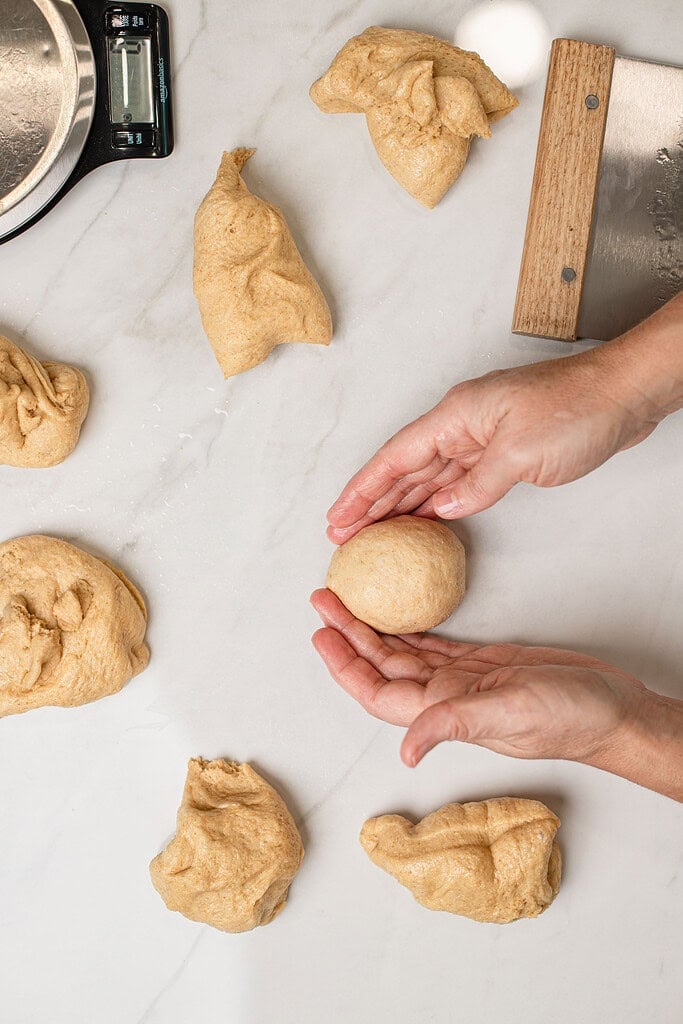 Overhead view of hands shaping a portioned piece of dinner roll dough into a smooth ball on a marble counter, with several other dough portions and a bench scraper and kitchen scale nearby.