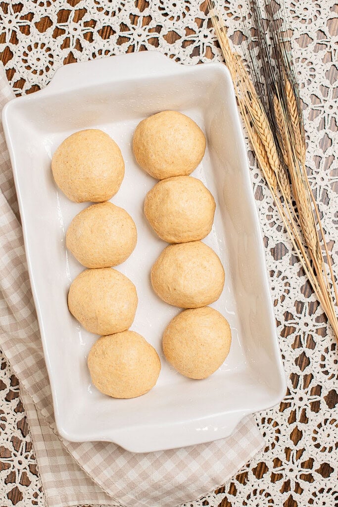Overhead view of eight shaped dinner roll dough balls arranged in rows in a white baking dish, not yet risen, with dried wheat stalks resting beside the pan.