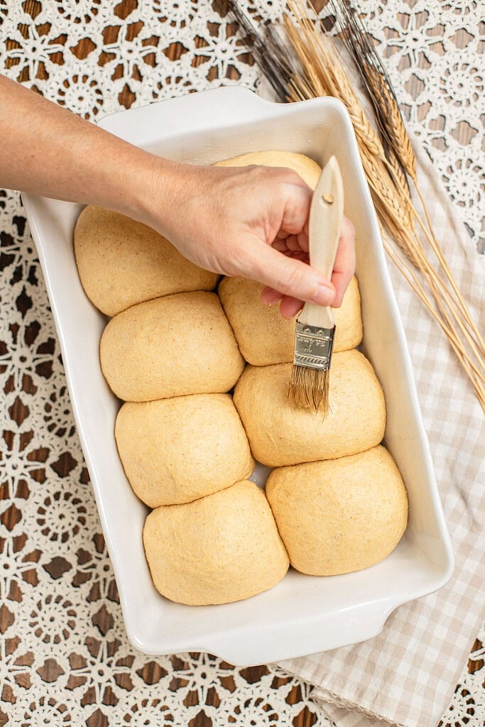 Overhead view of a hand brushing melted butter onto risen dinner rolls made with freshly milled flour in a white baking dish, with dried wheat stalks nearby.