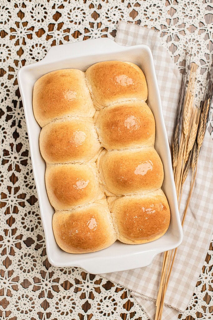Overhead view of golden brown baked dinner rolls made with freshly milled flour in a white baking dish, brushed with a glossy butter finish, with dried wheat stalks resting beside the pan.