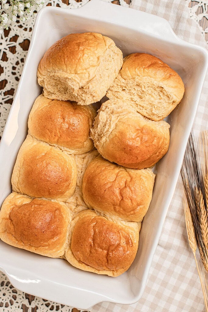 Overhead close-up of baked dinner rolls made with freshly milled flour in a white baking dish, one roll torn open to reveal the soft fluffy interior, with baby's breath flowers and dried wheat stalks in the background.