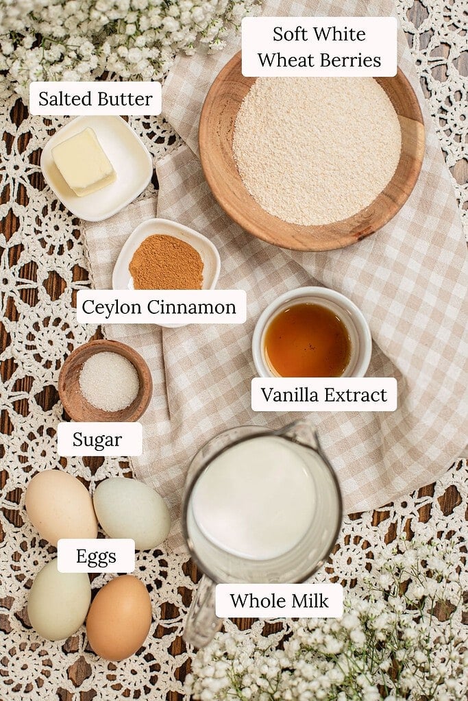 Flat lay of labeled ingredients for a freshly milled whole wheat Dutch baby pancake, including a bowl of soft white wheat berries and flour, salted butter, ground Ceylon cinnamon, vanilla extract, sugar, a pitcher of whole milk, and farm fresh eggs, arranged on a beige checked cloth with baby's breath flowers.