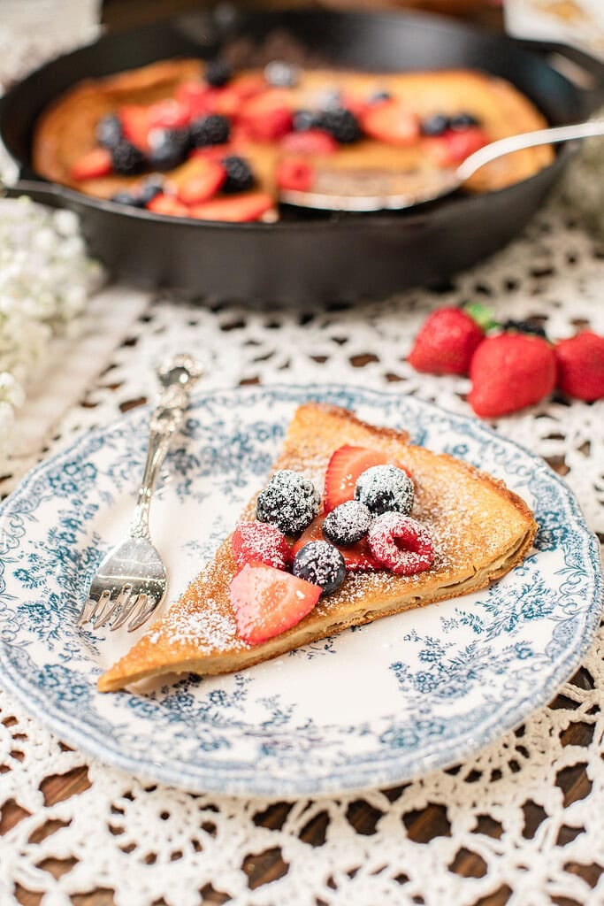 Slice of freshly milled whole wheat Dutch baby pancake on a blue and white floral plate, dusted with powdered sugar and topped with strawberries, blueberries, and blackberries, with a silver fork resting beside it and the cast iron skillet visible in the background.