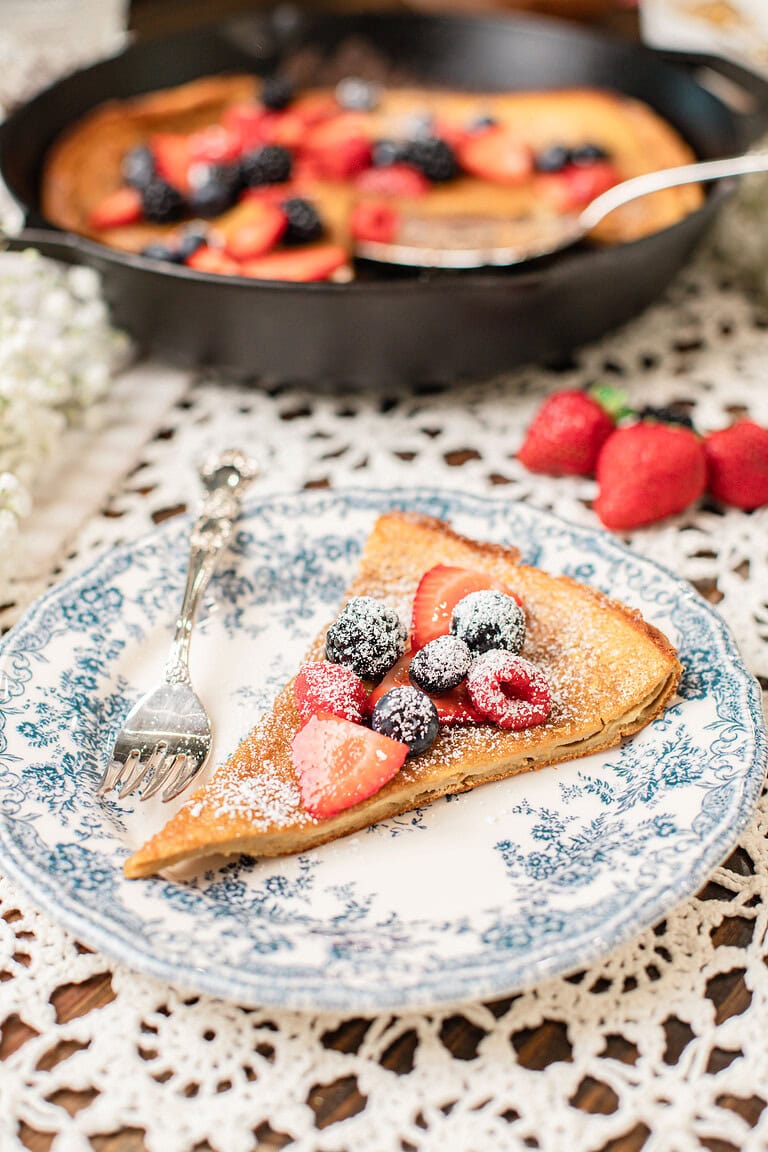 Slice of freshly milled whole wheat Dutch baby pancake on a blue and white floral plate, dusted with powdered sugar and topped with strawberries, blueberries, and blackberries, with a silver fork resting beside it and the cast iron skillet visible in the background.