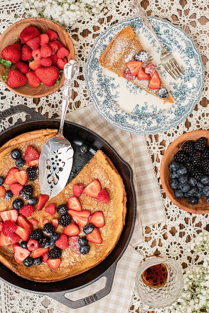 Overhead view of a soft white wheat Dutch baby in a cast iron skillet with a slice cut and lifted onto a floral plate, surrounded by wooden bowls of fresh strawberries, raspberries, and blackberries, and a small glass of maple syrup.
